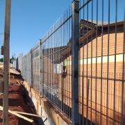 Foto externa, com uma obra em andamento na calçada e no muro ao lado da escola. Atrás da cerca é possível ver dois prédios da escola, de um piso e tijolo à vista. O céu está azul.