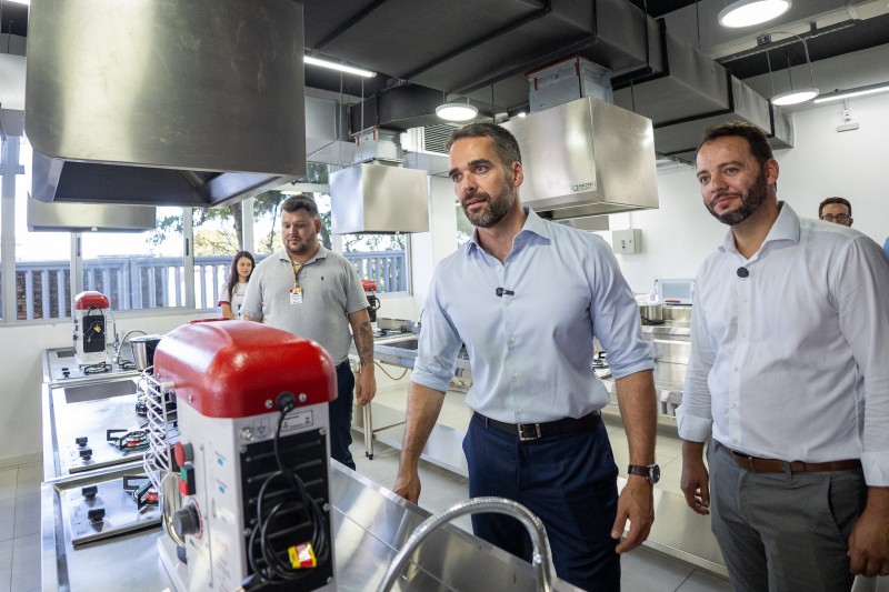 Em uma sala com equipamentos de culinária aparecem três homens.