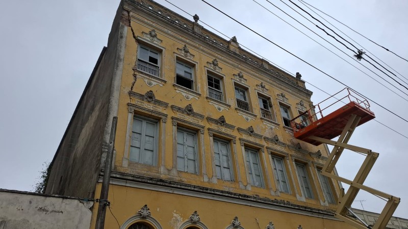 Foto de um prédio antigo de três andares com pintura amarela e rachaduras. Uma plataforma elevatória aparece à frente dele.