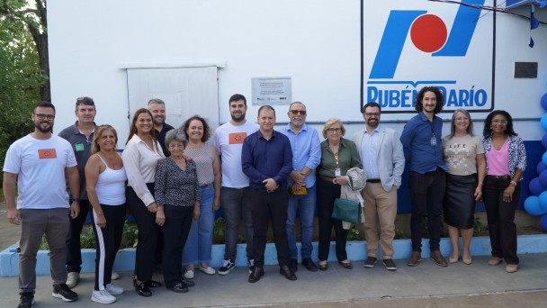 Grupo de pessoas posa em frente ao prédio da escola, onde há o logo da escola. Atrás deles há uma placa de metal.