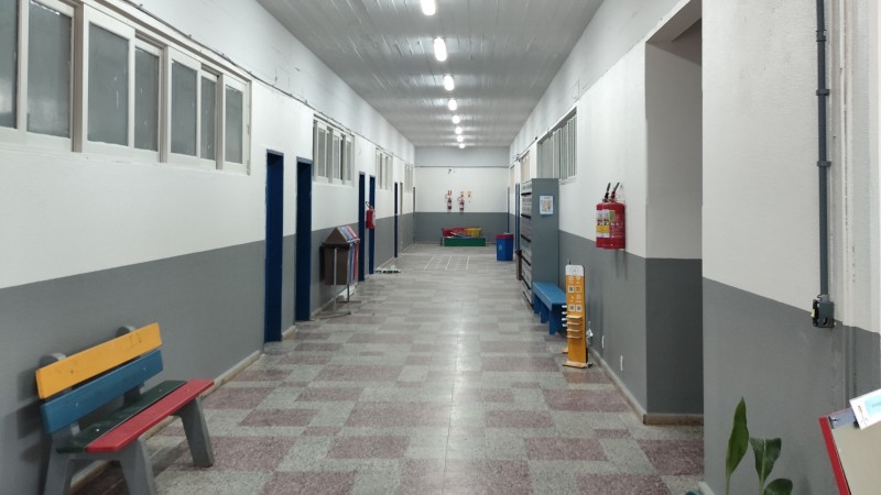 Foto interna de um corredor de escola pintado em cinza e branco, com as luzes acesas no teto.