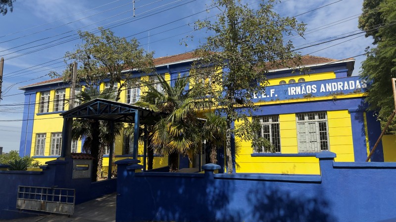 Foto da fachada da escola, um prédio de dois pisos pintado em azul e amarelo. À frente do prédio há árvores e um muro baixo, pintado de azul.