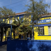 Foto da fachada da escola, um prédio de dois pisos pintado em azul e amarelo. À frente do prédio há árvores e um muro baixo, pintado de azul.
