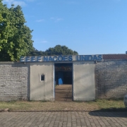 Foto da entrada de uma escola. Vê-se o muro branco da frente da escola, com o nome dela em um letreiro sobre a porta. Há um carro branco estacionado à direita.