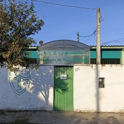 Foto da entrada de uma escola. Vê-se o muro branco e o letreiro com o nome da escola em cima da porta.
