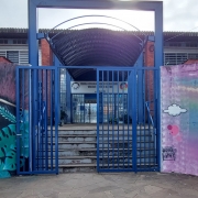 Foto da entrada da escola. Há pinturas coloridas no muro e a escola é um prédio de dois pisos.