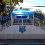 Foto da fachada da escola. Vê-se uma escadaria que leva ao alto de um morro onde está o prédio da escola. A escadaria tem uma cobertura azul. Há árvores em frente ao prédio e um letreiro à esquerda com o nome da escola.