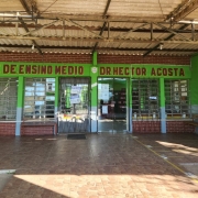 Foto da entrada da escola, um ambiente em U inverso pintado em verde com uma cobertura. Há lixeiras e um banco à direita e um banco e um quadro com frases à esquerda. Acima das portas, um letreiro indica o nome da escola.