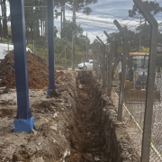 Foto do pátio da escola, onde se vê um buraco no chão ao centro, com a escola à esquerda e uma patrola do lado de fora da cerca, estacionada na rua. Ao fundo, veem-se dois carros circulando.
