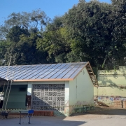 Foto do pátio da escola, com um prédio térreo à esquerda, pintado de verde e com o telhado retirado. Ao fundo, um muro alto verde é encimado por árvores e uma casa branca entre elas.
