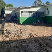 À esquerda uma casa verde. À direita e ao fundo o prédio térreo da escola, pintado em tons de verde. Em primeiro plano, terra mexida.