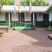 Em primeiro plano, vê-se um pátio de pedrinhas. Ao fundo e à esquerda, o prédio térreo da escola, pintado em branco e verde. Em frente à ele, à esquerda, uma árvore e, no centro, quatro bandeiras em postes. Há materiais de construção espalhado no terreno.