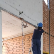 Um homem vestido de azul e usando capacete de obra azul mexe na calha de fios elétricos presa em uma parede de tijolos à vista.