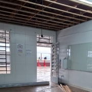 De dentro de uma sala pintada de branco, vê-se ao fundo o pátio da escola. A porta está aberta. O forro do telhado foi retirado e há um quadro de vidro à direita.