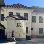 Em frente ao prédio de dois pisos pintado de branco, um homem de uniforme e capacete de obra passa carregando uma estrutura de metal. Há uma cesta de basquete na parede da escola.