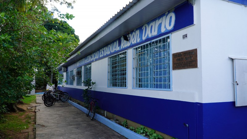 Foto do prédio térreo da escola, pintado em branco e azul, com o nome pintado na parede e uma placa presa. Há bicicletas em frente ao prédio.