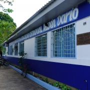 Foto do prédio térreo da escola, pintado em branco e azul, com o nome pintado na parede e uma placa presa. Há bicicletas em frente ao prédio.