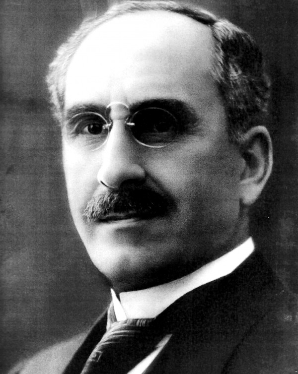 Retrato em preto e branco de um homem branco de bigodes, usando óculos e terno e gravata.