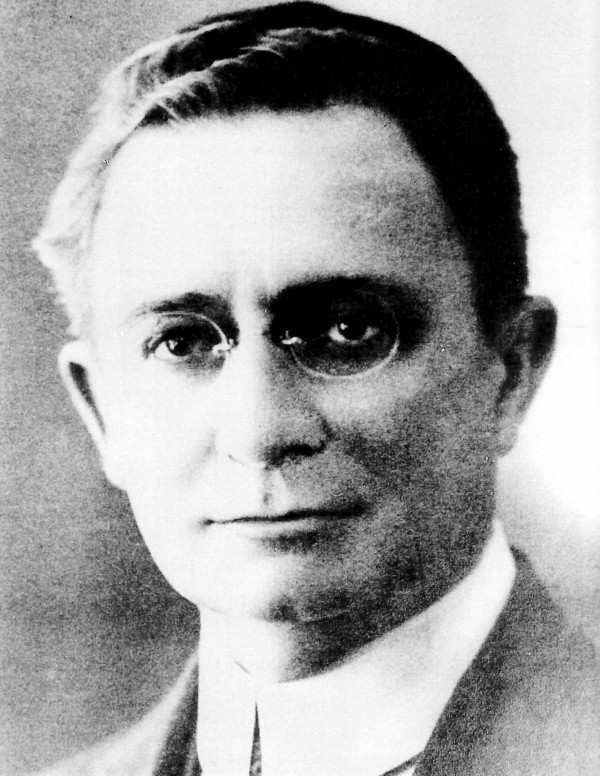 Foto em preto e branco de um homem branco usando óculos e vestindo terno e gravata.