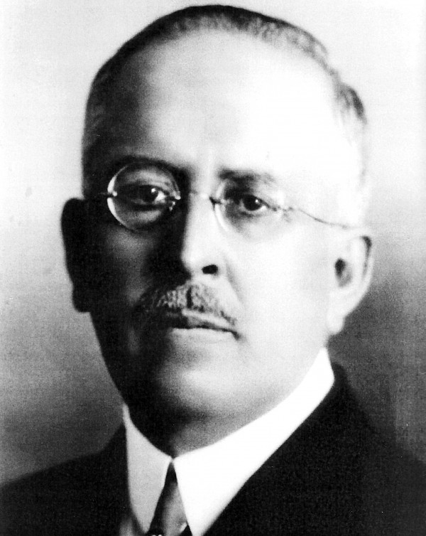 Retrato em preto e branco de um homem branco de bigode, usando óculos e terno e gravata.