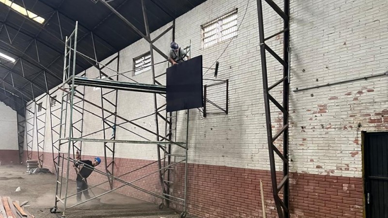 Dentro de um ginásio, um homem em cima de um andaime instala uma tabela de basquete na parede. Há outro homem no chão, varrendo.
