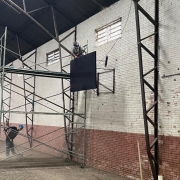 Dentro de um ginásio, um homem em cima de um andaime instala uma tabela de basquete na parede. Há outro homem no chão, varrendo.