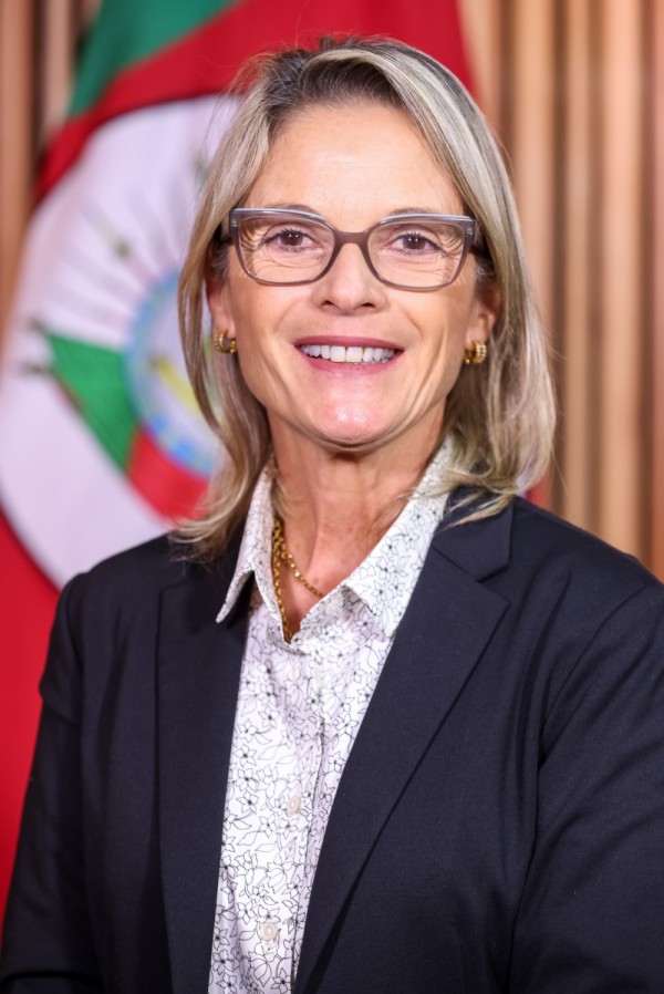 Foto colorida de uma mulher loira sorridente de óculos. Ao fundo, desfocada, se vê a bandeira do Rio Grande do Sul.