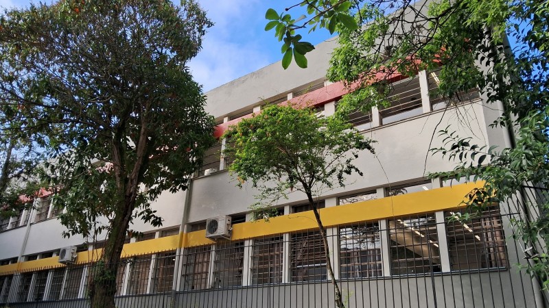 Foto de um prédio de escola de dois pisos, pintado em cinza e com detalhes em amarelo e vermelho. Há árvores à frente e o céu está azul.