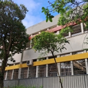 Foto de um prédio de escola de dois pisos, pintado em cinza e com detalhes em amarelo e vermelho. Há árvores à frente e o céu está azul.
