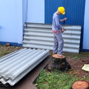 Um homem de capacete de obra amarelo segura uma corda. Atrás dele, o prédio da escola, pintado em tons de azul. Há telhas espalhadas pelo pátio.