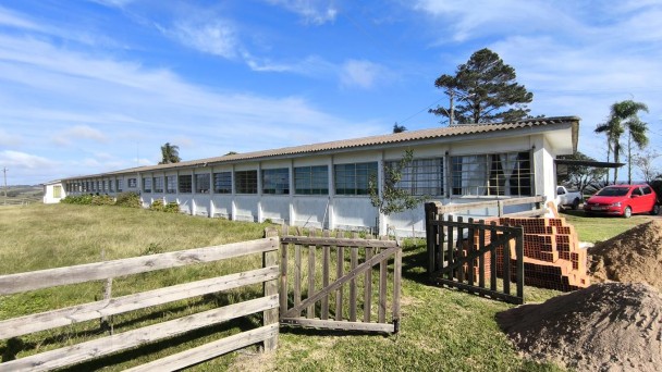 Foto do prédio da escola, de um piso, pintado em branco. Em primeiro plano, uma cerca baixa de madeira, com o portão aberto, um gramado e materiais de construção. Há carros próximo da escola e o céu está azul.