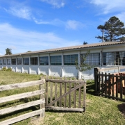 Foto do prédio da escola, de um piso, pintado em branco. Em primeiro plano, uma cerca baixa de madeira, com o portão aberto, um gramado e materiais de construção. Há carros próximo da escola e o céu está azul.