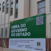 Foto da fachada da escola, um prédio de dois pisos pintado de bege. Em primeiro plano vê-se a placa de obra e a cerca.