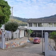 Foto do prédio da escola, de dois pisos pintado em cinza. Vê-se, em primeiro plano, o pátio da escola, com carros estacionados. À esquerda, outro prédio da escola, térreo, com um parquinho infantil e uma árvore à frente. No fundo, morros e o céu com nuvens.