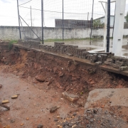 Foto de uma área em obras. Num nível mais baixo, em primeiro plano, vê-se uma calçada com barro. Há um desnível de onde foram retirados os tijolos que compunham o muro. No nível mais alto, uma quadra de esportes descoberta. O tempo está chuvoso.