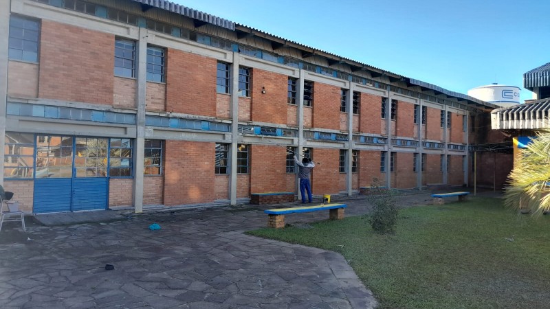 Foto do pátio interno da escola, com o prédio de dois pisos de tijolo à vista da escola ao fundo. Um homem pinta a parede. Ao fundo, se vê o céu azul e uma caixa d'água da Corsan.