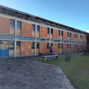 Foto do pátio interno da escola, com o prédio de dois pisos de tijolo à vista da escola ao fundo. Um homem pinta a parede. Ao fundo, se vê o céu azul e uma caixa d'água da Corsan.