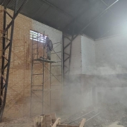 No interior de um ginásio, em um ambiente enfumaçado, um homem está parado em um andaime. Há entulhos no piso. As paredes estão em tijolo à vista.
