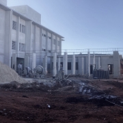 Foto de uma obra em andamento. À esquerda, um prédio de três pisos sem pintura. Ao centro, o início de uma construção. Vê-se homens trabalhando e materiais de construção no pátio.