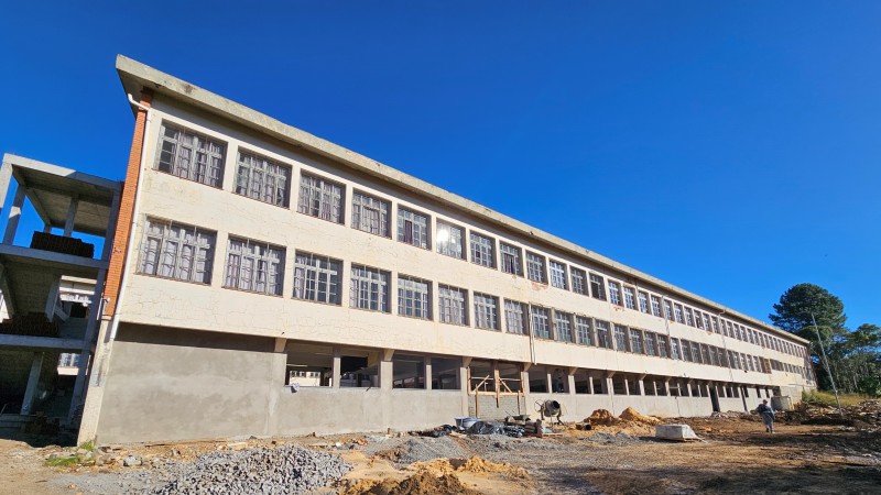 Foto de um prédio em construção, com três pisos. Há um homem no pátio, onde também se veem materiais de construção. O céu está azul.