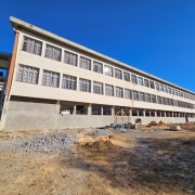 Foto de um prédio em construção, com três pisos. Há um homem no pátio, onde também se veem materiais de construção. O céu está azul.