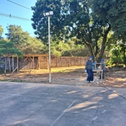 No pátio da escola, veem-se três homens de capacete de obra. Há árvores e um poste.