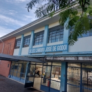 Foto da fachada da escola, um prédio de dois pisos, com parte em tijolo a vista e parte pintado de branco e azul. Há um letreiro na fachada com o nome da escola.