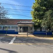 Foto do prédio da escola visto do outro lado da rua. Em primeiro plano, a rua. Após, o prédio de um piso da escola, com um toldo no corredor de entrada e árvores à frente.