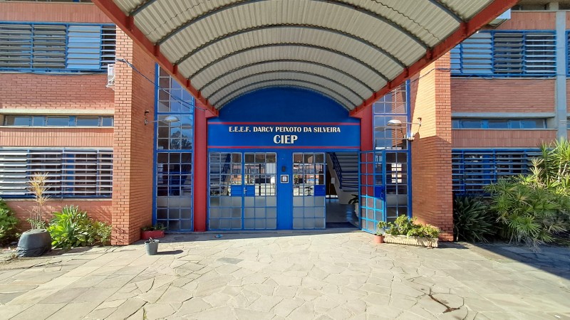 Foto da entrada da escola. O prédio tem dois pisos e é de tijolo à vista. A porta é azul e tem, sobre ela, um letreiro com o nome da escola. Há uma cobertura na calçada que leva à entrada.