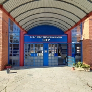 Foto da entrada da escola. O prédio tem dois pisos e é de tijolo à vista. A porta é azul e tem, sobre ela, um letreiro com o nome da escola. Há uma cobertura na calçada que leva à entrada.