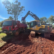 Em uma área de grama e terra vermelha, uma retroescavadeira joga terra na caçamba de um caminhão vermelho. Há árvores ao fundo e o céu está azul.