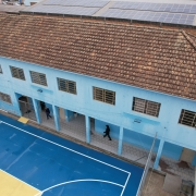 Foto aérea mostra o prédio de dois pisos da escola, pintado em azul claro. É possível ver parte da quadra de esportes descoberta, pintada em azul e amarelo.