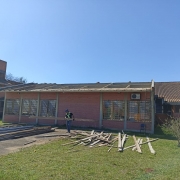 Em frente a um prédio térreo de um piso da escola, em tijolo à vista, um homem carrega uma madeira. O telhado está parcialmente retirado. Há outras madeiras pelo chão. Ao fundo, há outro prédio de tijolos à vista, de três pisos, no canto. O céu está azul.