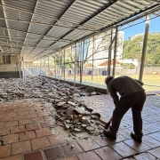 Em uma área coberta, um homem de capacete quebra um piso. Ao fundo se vê uma quadra de esportes descoberta.